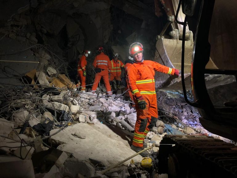 Men in orange jumpsuits, with head torches, search through rubble at night. They are members of the UK's International Search & Rescue team, in Hatay, Turkey, looking for survivors of the 2023 earthquake.