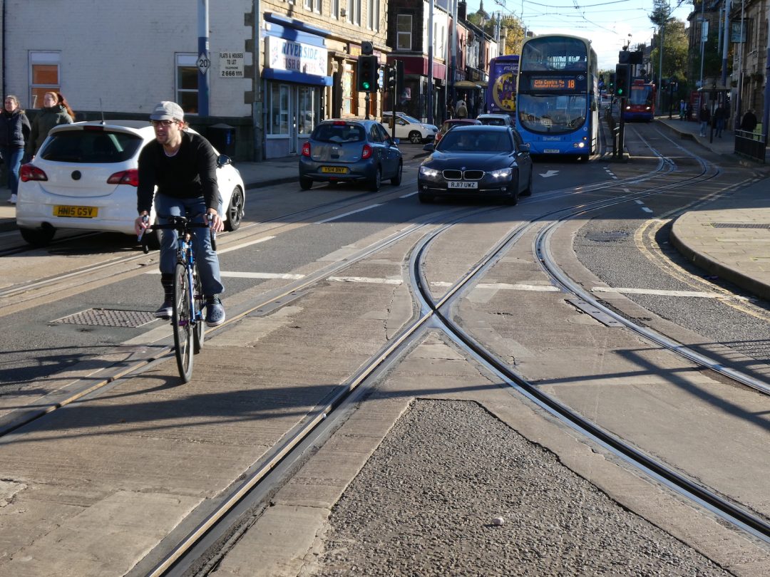 A cyclist riding infront of a car and buses on a road with tramrail tracks.