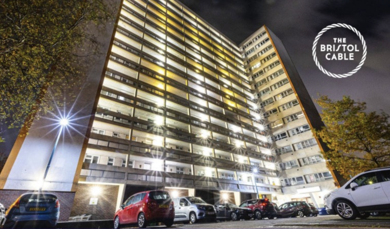 A white-painted tower block at night, with cars parked at the base.