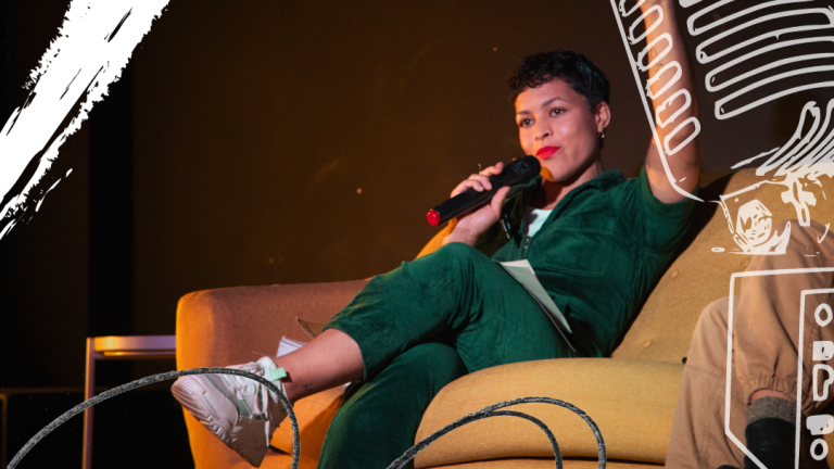 A person with a microphone sat on a sofa wearing trainers and a velvet jumpsuit.