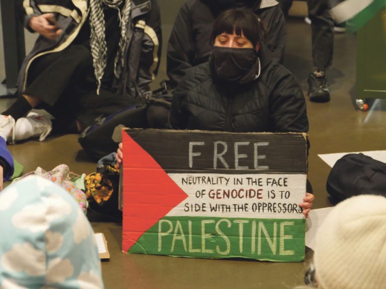 A woman with a face covering sits behind a placard marked 'Free Palestine'