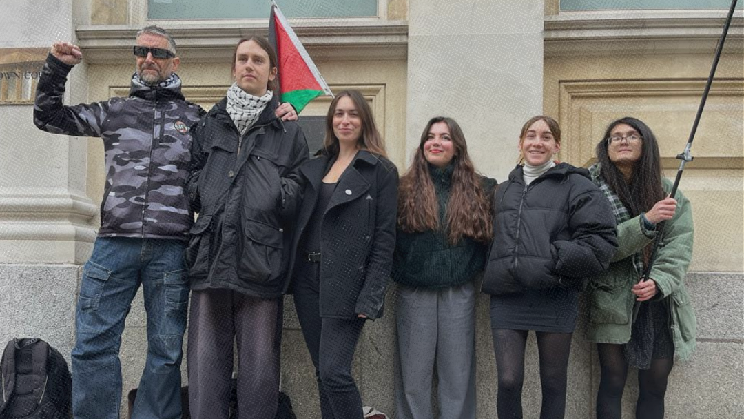 Seven people stood outside a building. Two are holding Palestine flags.