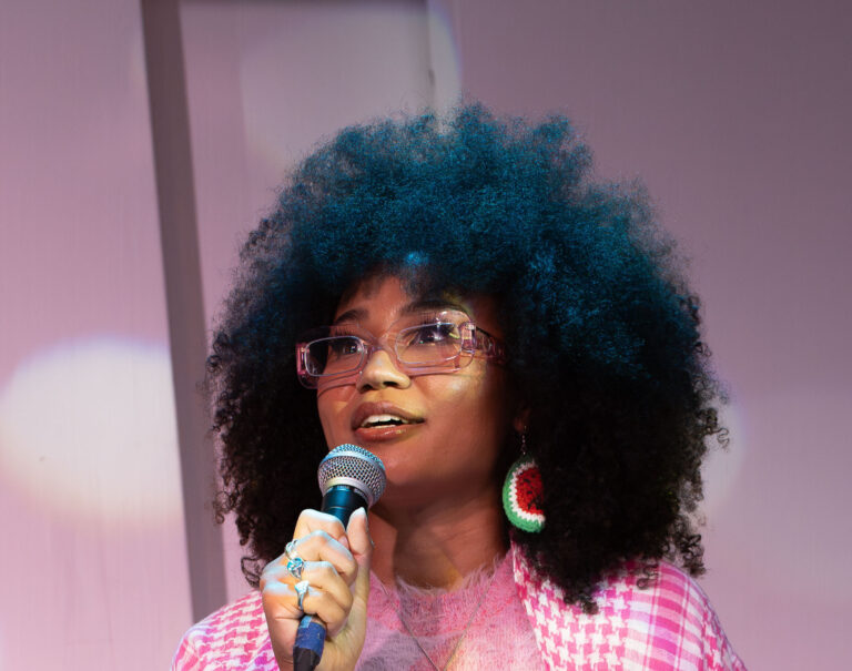 A woman with dressed in pink and wearing glasses speaks into a microphone