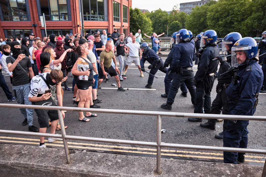 A crowd of far-right protesters are standing off against a line of police officers. One office is wielding a baton.