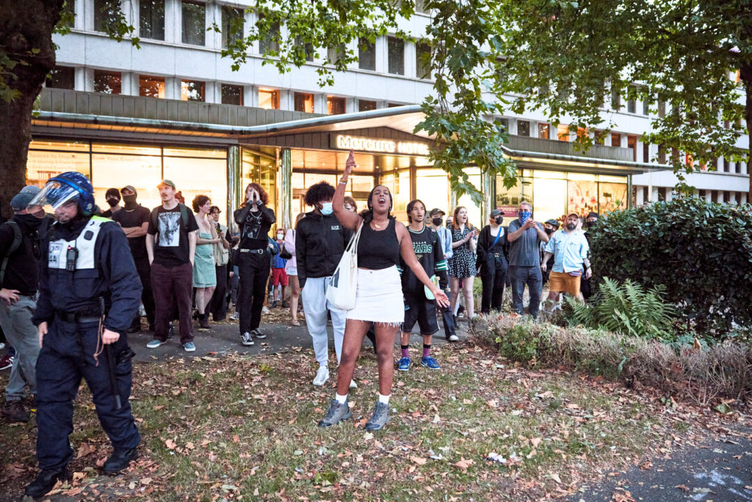 A group of anti-fascists are stood chanting outside a hotel. 