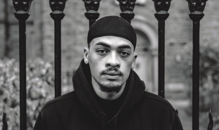 A man in a black hoodie and hat, seen in head-and-shoulders view, stands against a set of ornate railings