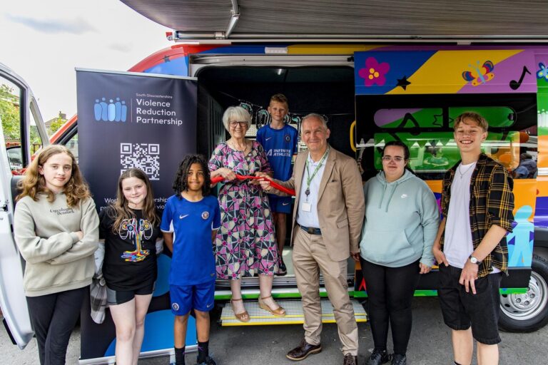 youth bus opening ceremony, several young people gathered around two adults who are about to cut a read ribbon all in front of a colourful bus.