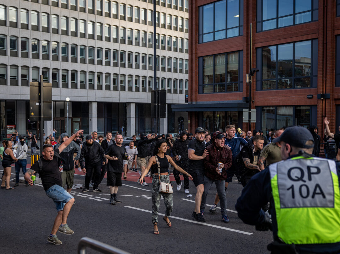 A group of protesters shout aggressively towards police. Some of them have cans of beer and cider in their hands. 