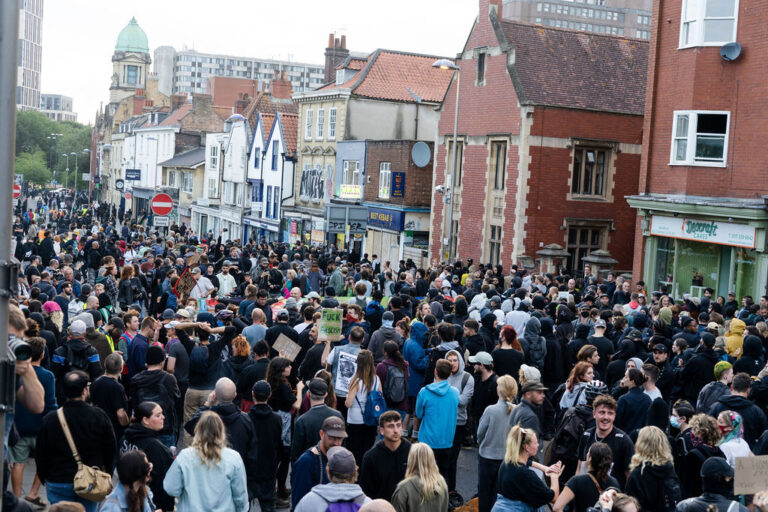 A large crowd gather in the road on a city street.