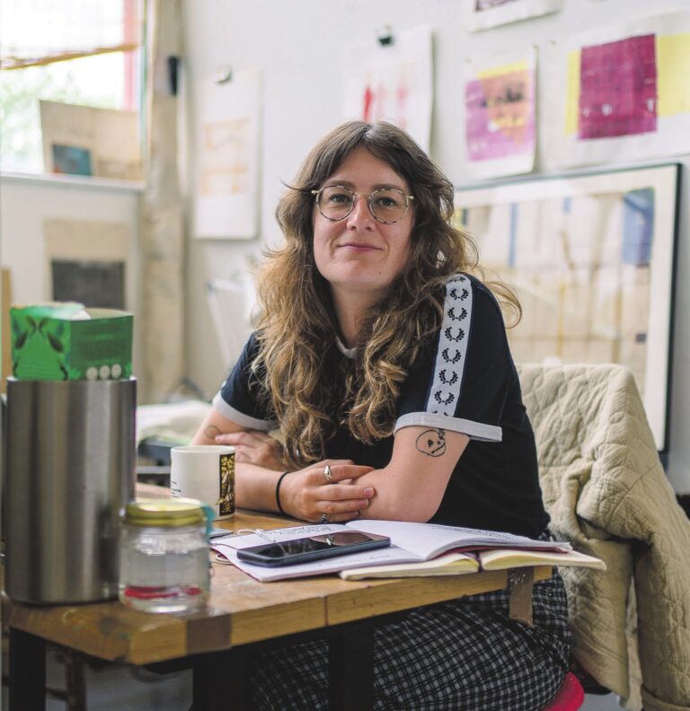 A women sits at a desk. On the wall behind are various art works.