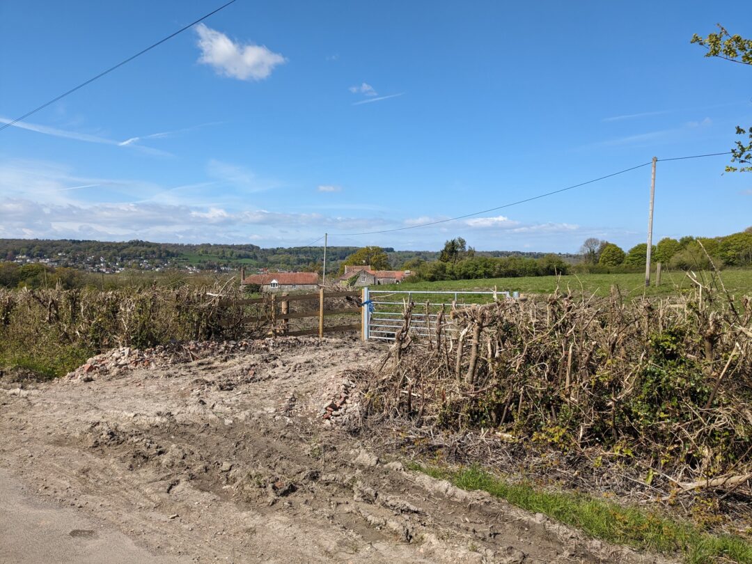 A damaged hedgerow on either side of a metal gate, with buildings in the distance.