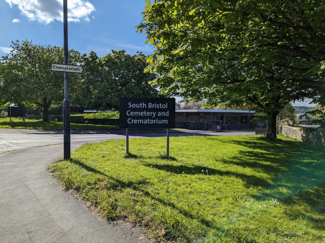 A sign for South Bristol Cemetery and Crematorium on a grass lawn next to a tree.