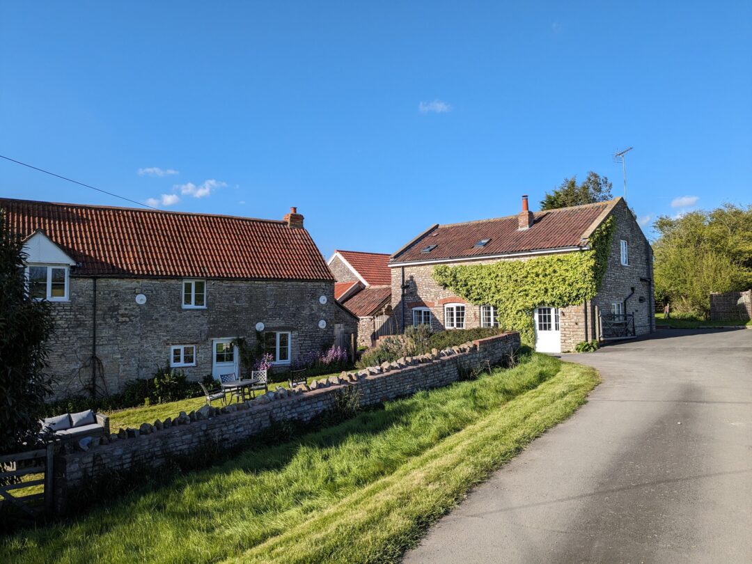 Two farmhouse buildings with a stone wall along a road.