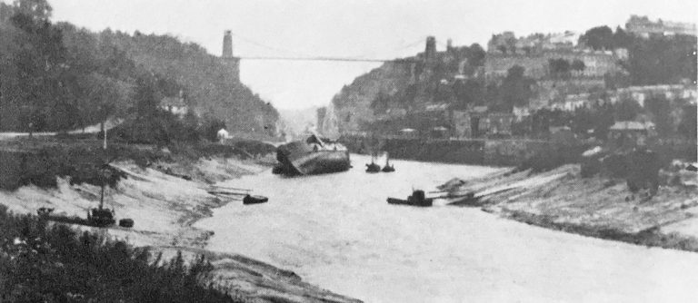 A black and white photo of the River Avon with a steam boat listing to the left framed by the Clifton Suspension Bridge.