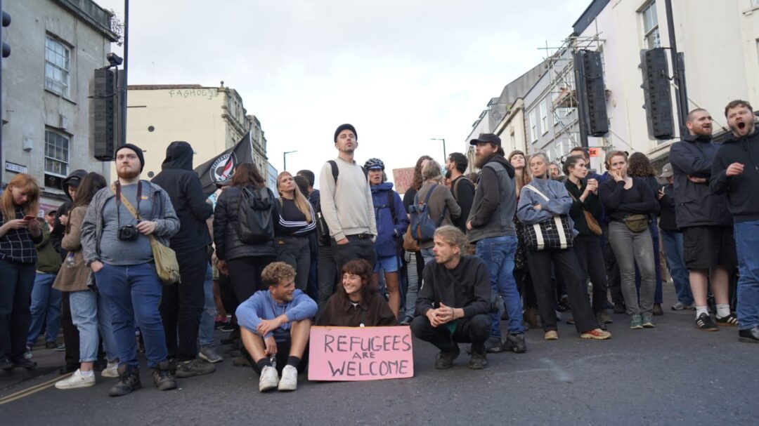 A crowd of protestors stand and sit in the street behind a placard saying 'refugees are welcome'