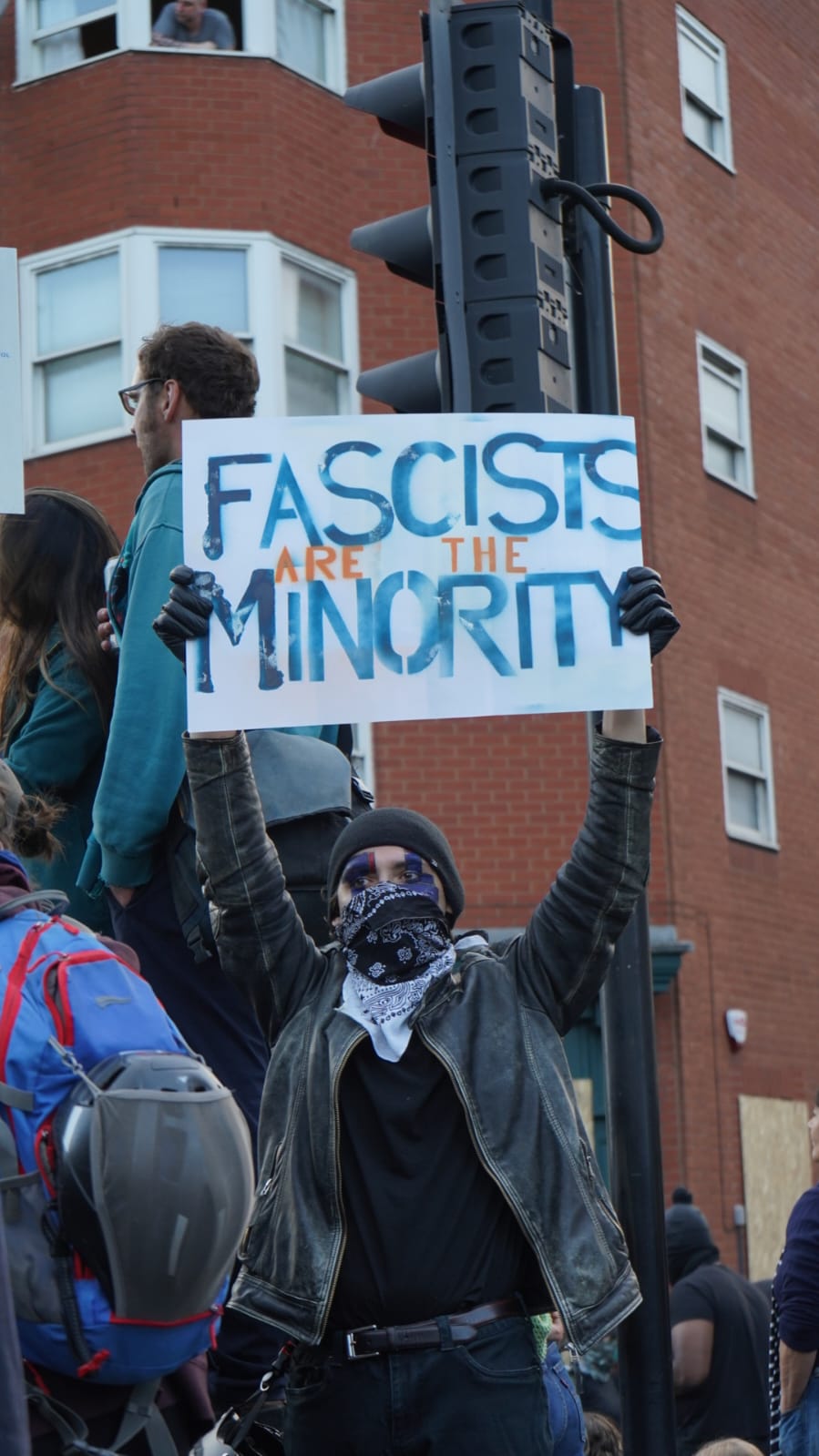 A protestor with sunglasses and a balaclava holds a placard saying 'fascists are the minority'