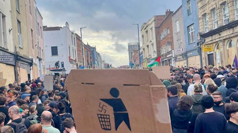 A cardboard placard showing a swastika being thrown in a bin is raised above a city street full of people protesting