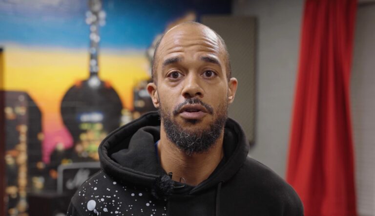 A man in a dark hoodie standing in a room with a mural of a cityscape in the background.