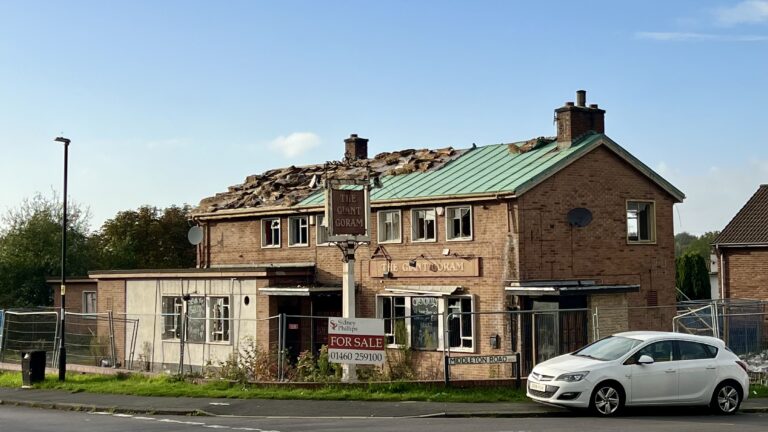 Exterior view of the Giant Goram pub with significant roof damage. A 'For Sale' sign is displayed in front of the building, with a parked car nearby.