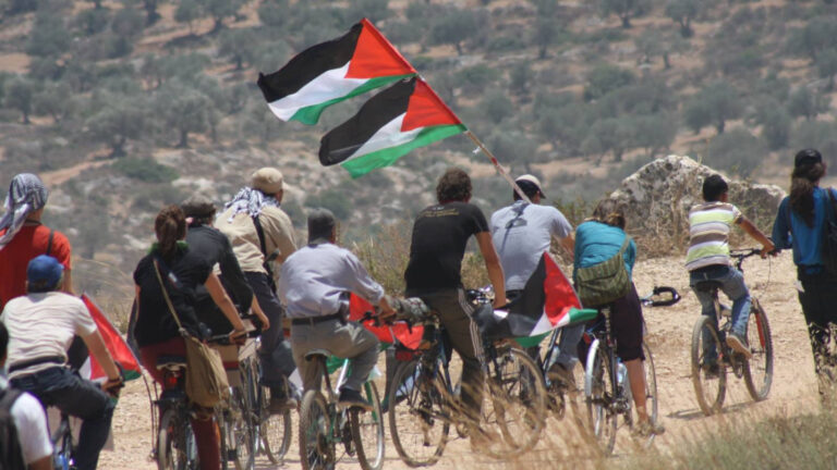 A group of individuals riding bicycles in a rural landscape, waving Palestinian flags.