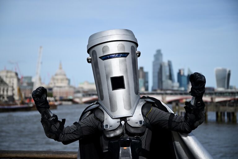 A person in a robotic costume with raised fists, standing on a bridge with the London skyline including St. Paul's Cathedral in the background.