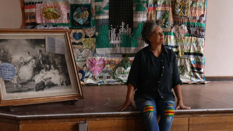 A women sitting on a wooden stage beside a quilt and a framed historic photograph.