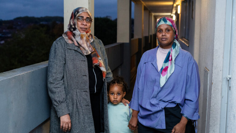 Two adults and a young child stand on a high-rise walkway with a twilight sky in the background. Both adults are wearing colourful headscarves.