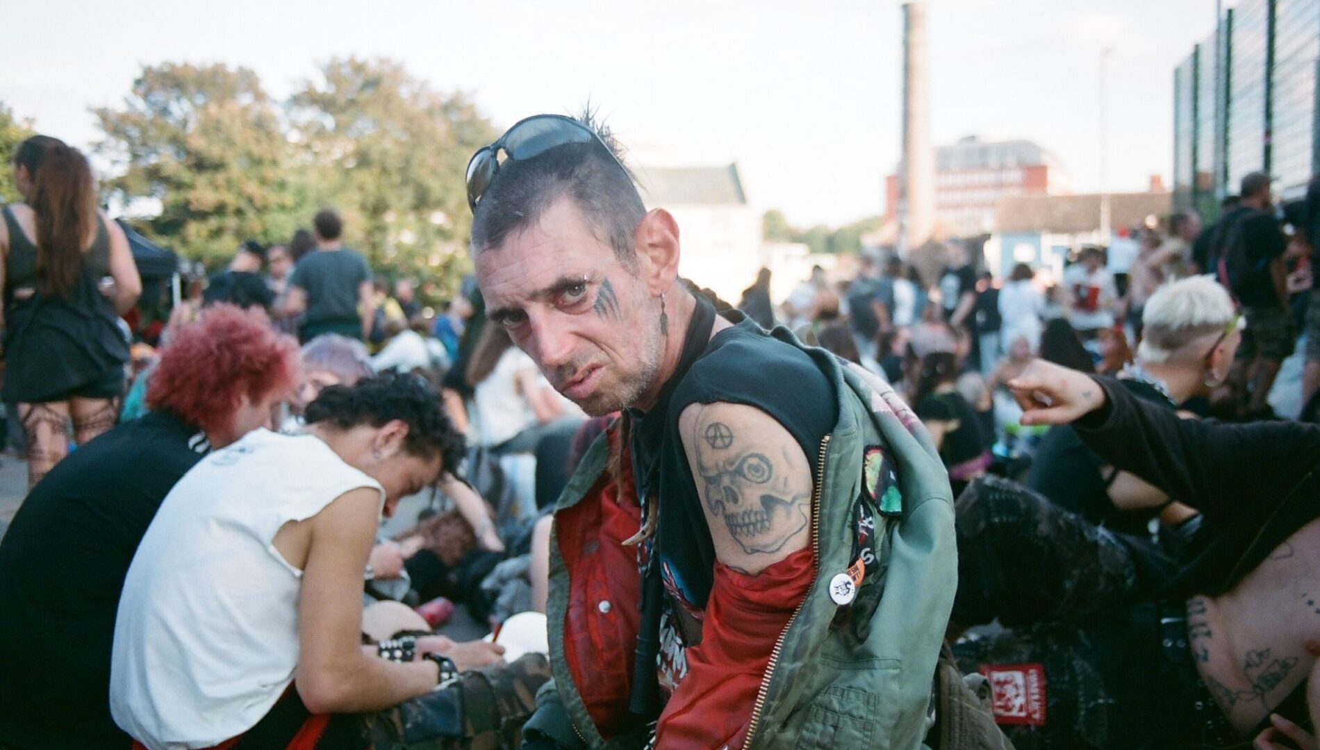 A person with a mohawk and tattooed arms is sitting outdoors at what appears to be a festival gathering. The setting includes other casually dressed individuals in a relaxed, social atmosphere.