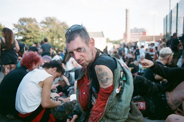 A person with a mohawk and tattooed arms is sitting outdoors at what appears to be a festival gathering. The setting includes other casually dressed individuals in a relaxed, social atmosphere.