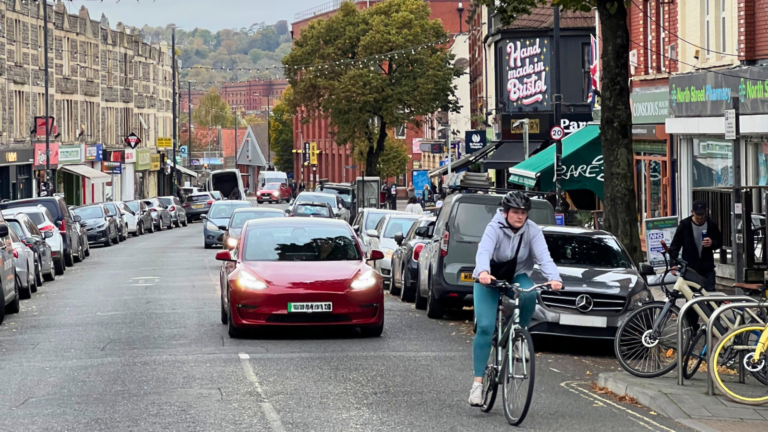 Urban street scene showing a cyclist in the foreground and a lineup of parked cars on either side, with commercial buildings.