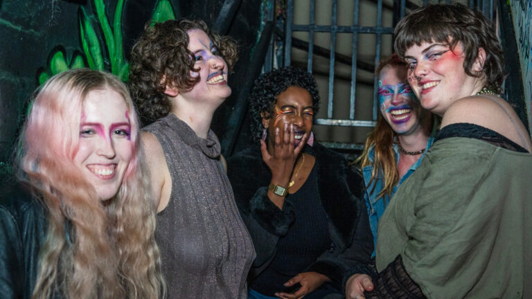 Group of five joyful people wearing bright makeup, laughing together in front of a graffiti-covered wall at night.