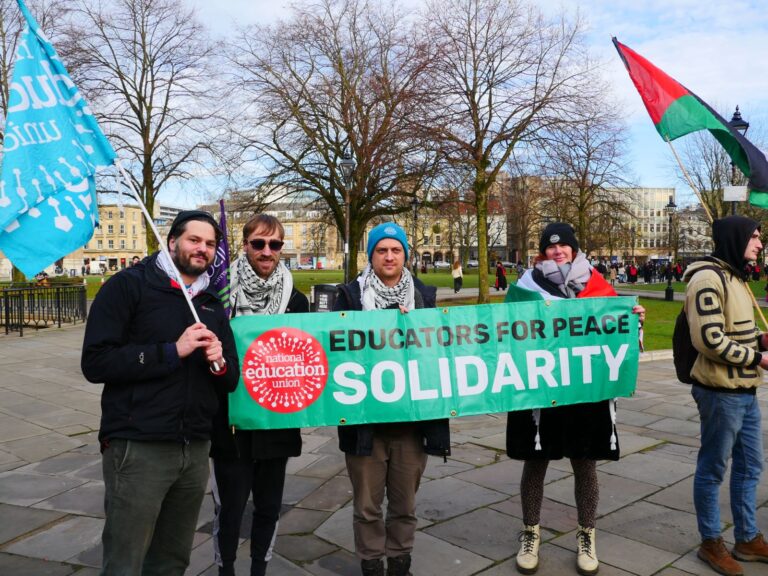 Four individuals holding a banner that reads 'Educators for Peace: Solidarity' at an outdoor rally, with union and Palestinian flags visible, and a park setting in the background.