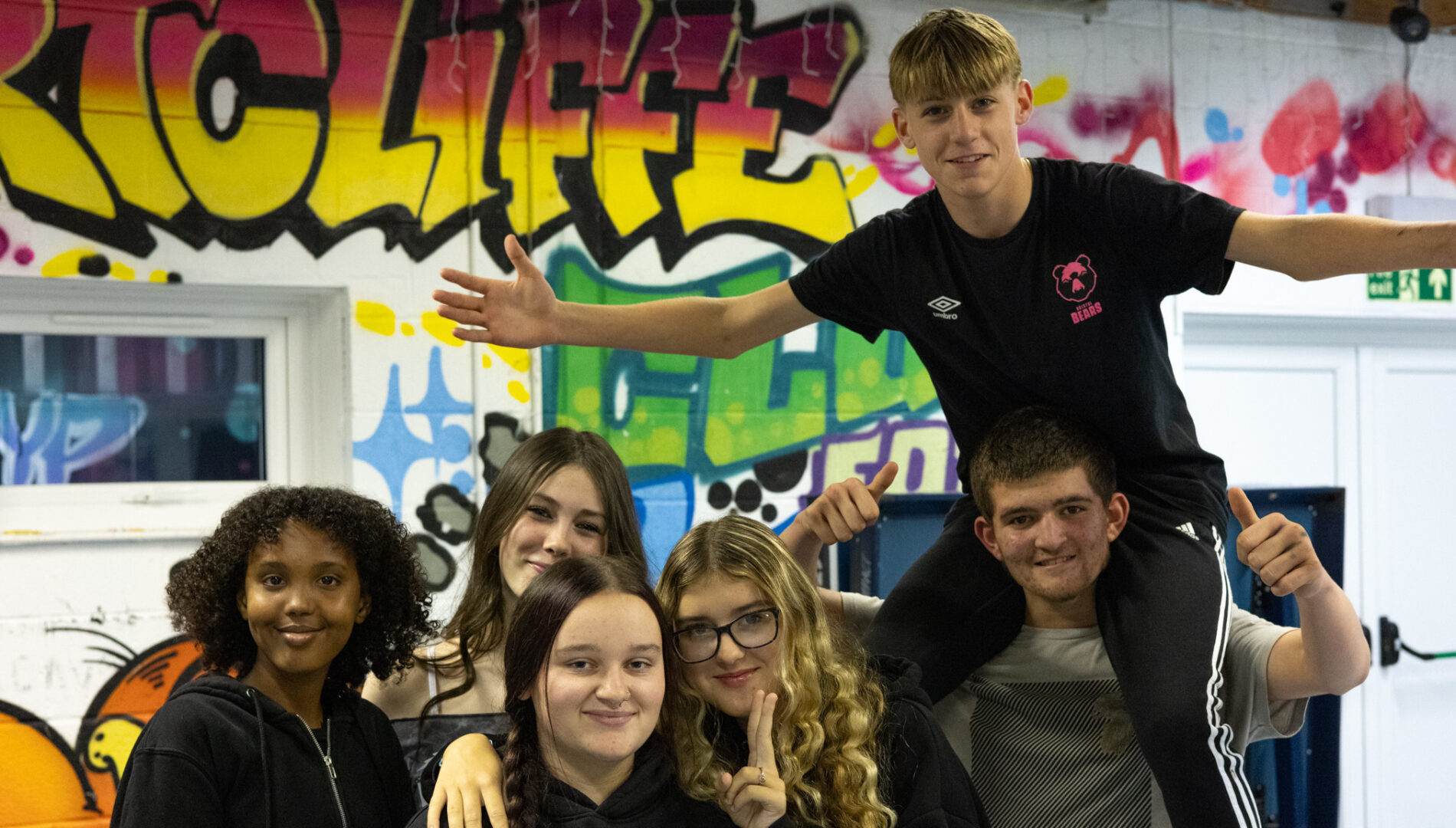 Group of six cheerful young people posing together in front of a colorful graffiti wall with the word 'Hartcliffe' (partly obscured) on it.