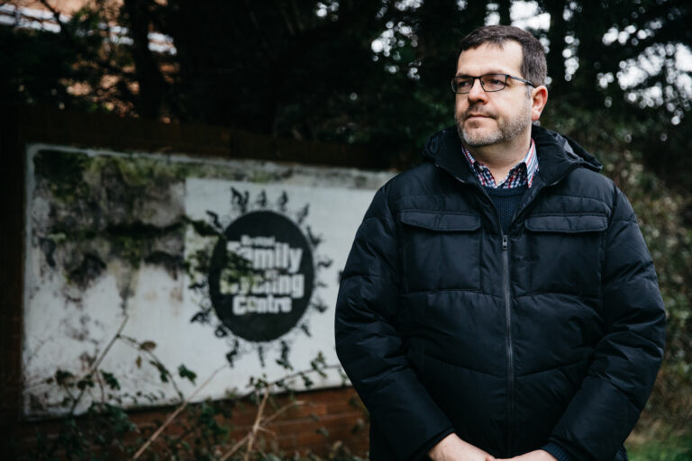 A man in a dark jacket stands in front of an overgrown Family Caring Centre sign, surrounded by trees and branches.