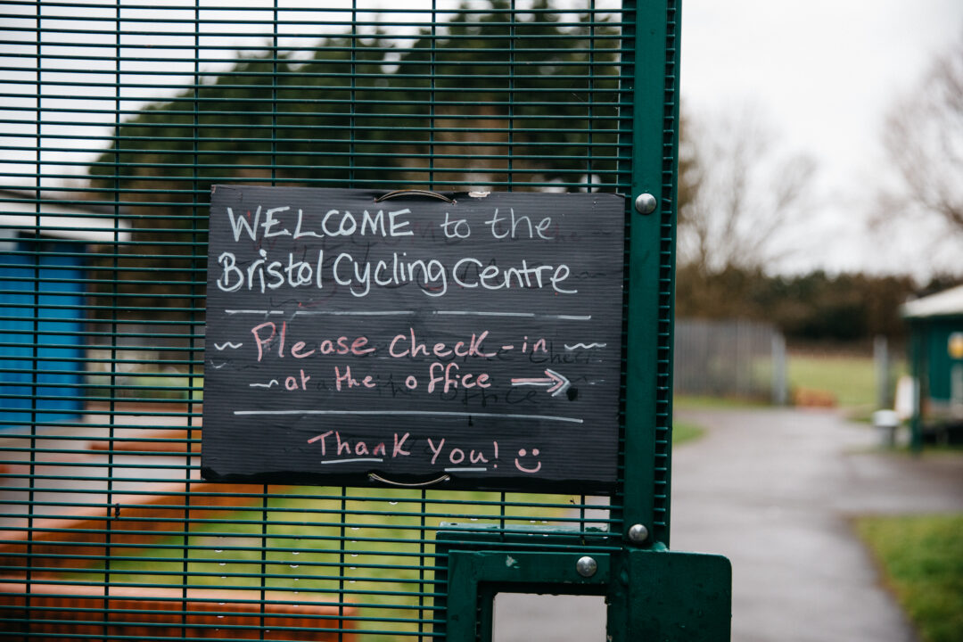 Sign on a metal gate reads, "Welcome to the Bristol Cycling Centre. Please check-in at the office. Thank you!" Path and greenery visible in the background.