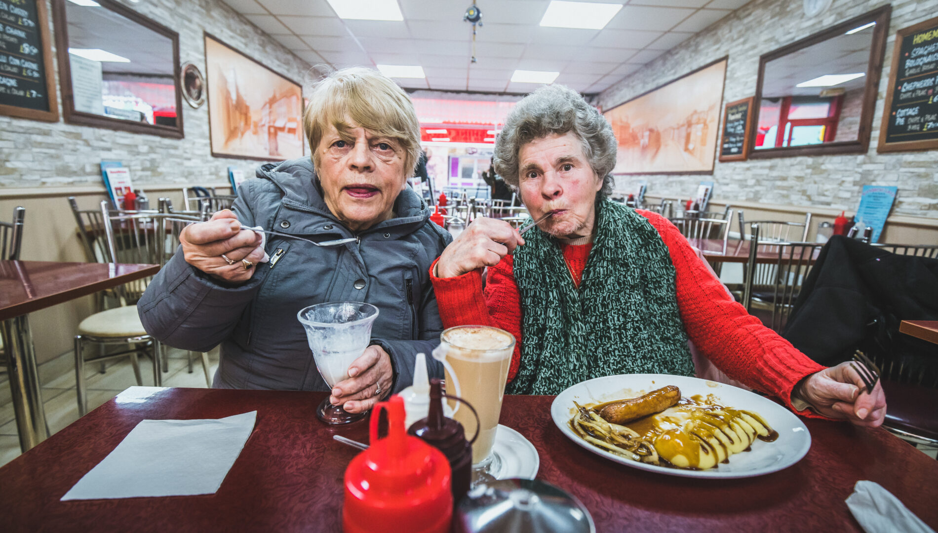 Two older women enjoy ice cream and sausage and mash together, at the table of a cafe.