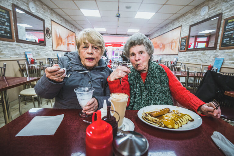 Two older women enjoy ice cream and sausage and mash together, at the table of a cafe.