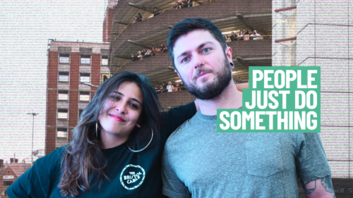 Two individuals smiling at the camera, standing close together. One is wearing a 'Bristol Cable' branded T-shirt and the other a plain green T-shirt. The text 'PEOPLE JUST DO SOMETHING' is displayed across the front. In the background is a grainy image of high-rise buildings.