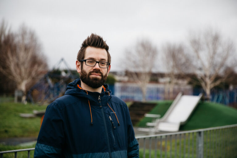 A man with glasses and a beard stands in front of a park playground, showcasing a casual yet thoughtful demeanour.