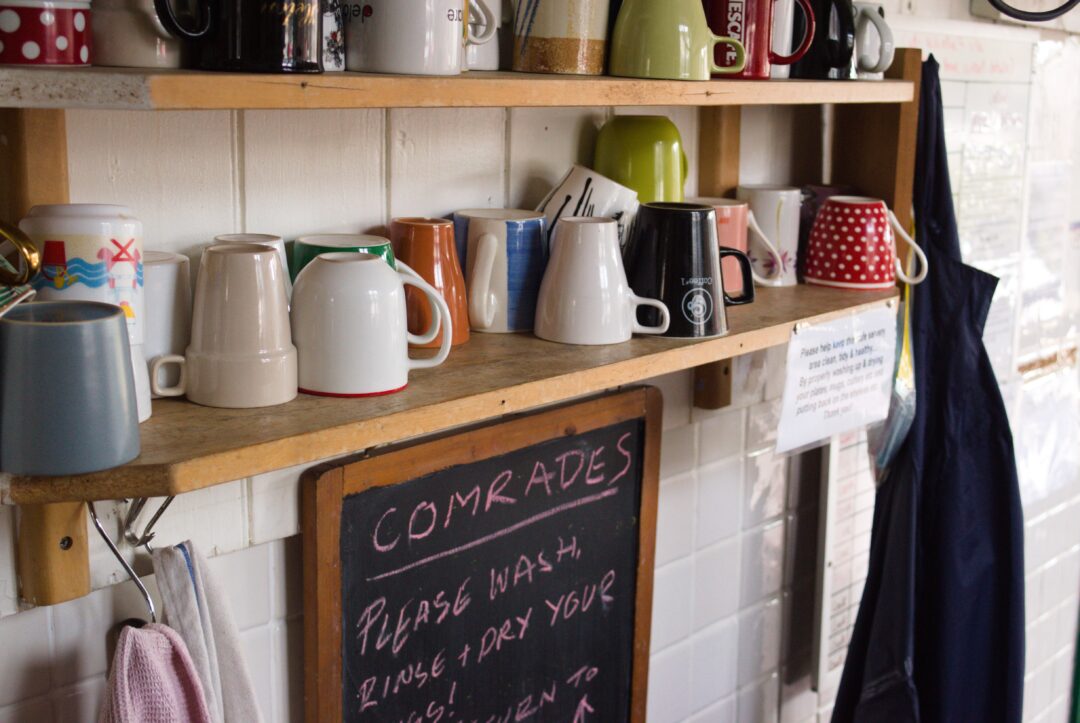 A row of cups on a wooden shelf with a black chalkboard sign below.