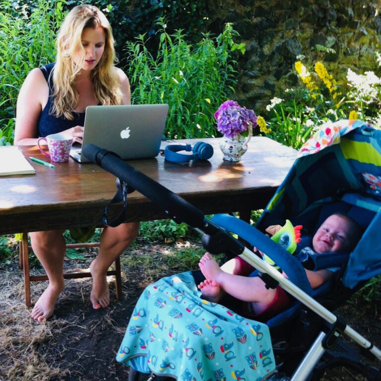 A woman with blonde hair works on her laptop at a table in her garden with her child in a pushchair in the foreground