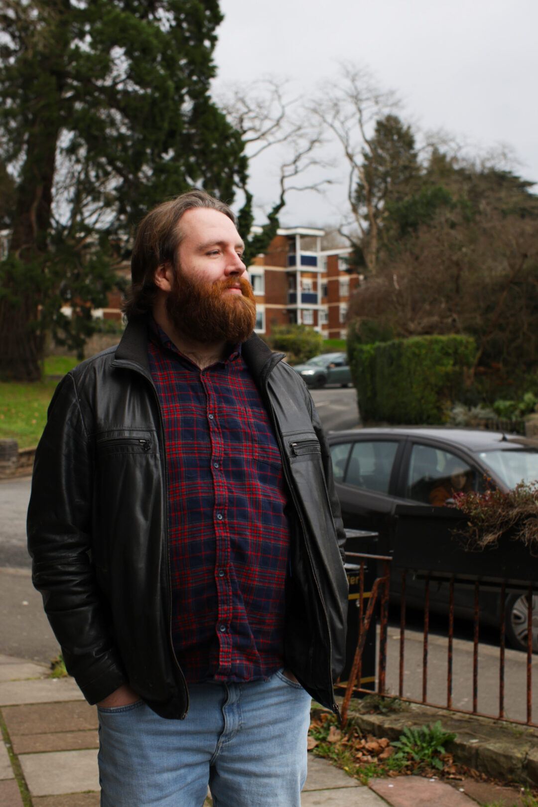 A man with a ginger beard, a black leather jacket and a red and blue checked shirt stands on the pavement with a small block of flats in the distance.