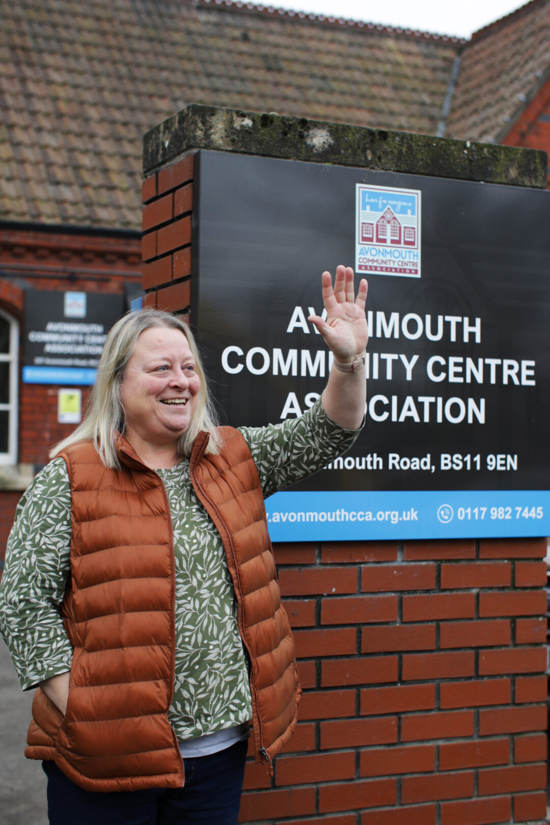 A woman wearing an orange gilet smiles and waves at the camera in front of the Ammouth Community Centre.
