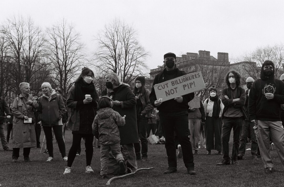 A protester holds a sign saying 'cut billionaires not PIP' in a crowd of other protesters standing on grass.