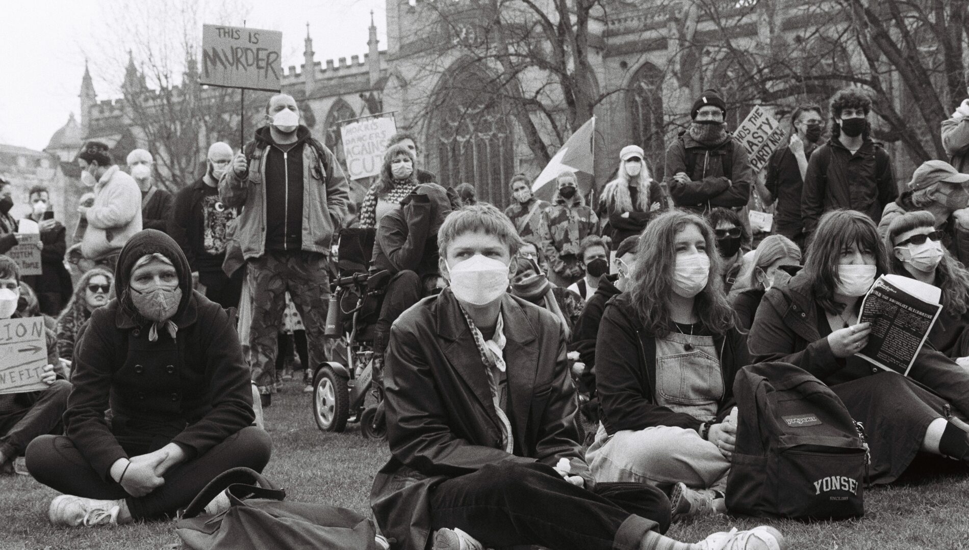 A group of protesters, wearing protective masks, some standing holding banners and some sitting on the grass in front of a cathedral.