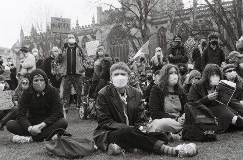 A group of protesters, wearing protective masks, some standing holding banners and some sitting on the grass in front of a cathedral.