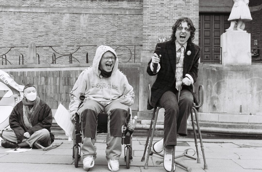 Three people sat on stage at a protest. One person is cross legged, while the person in the middle sits in a wheel chair and is smiling, next to a person wearing a dark jacket, also smiling.