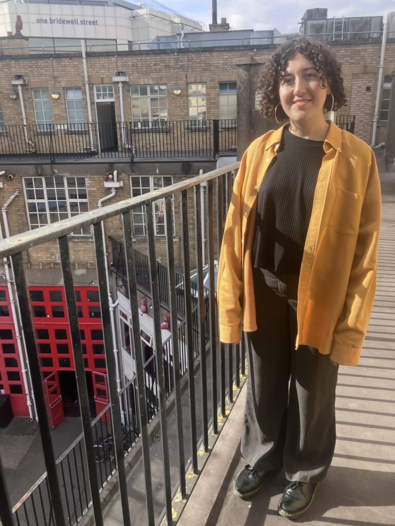 A woman and standing and smiling on a balcony, wearing a yellow jacket and black trousers. A bright red tour is on the ground floor.