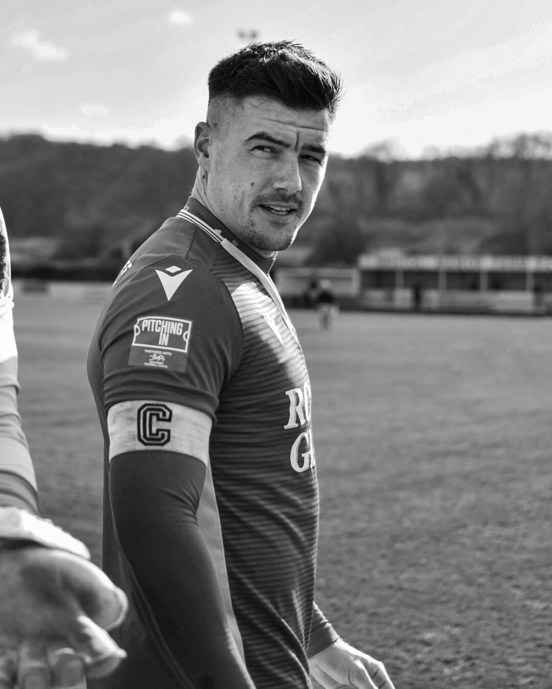 A football player in a long-sleeved jersey stands on a football pitch, with stands in the background.