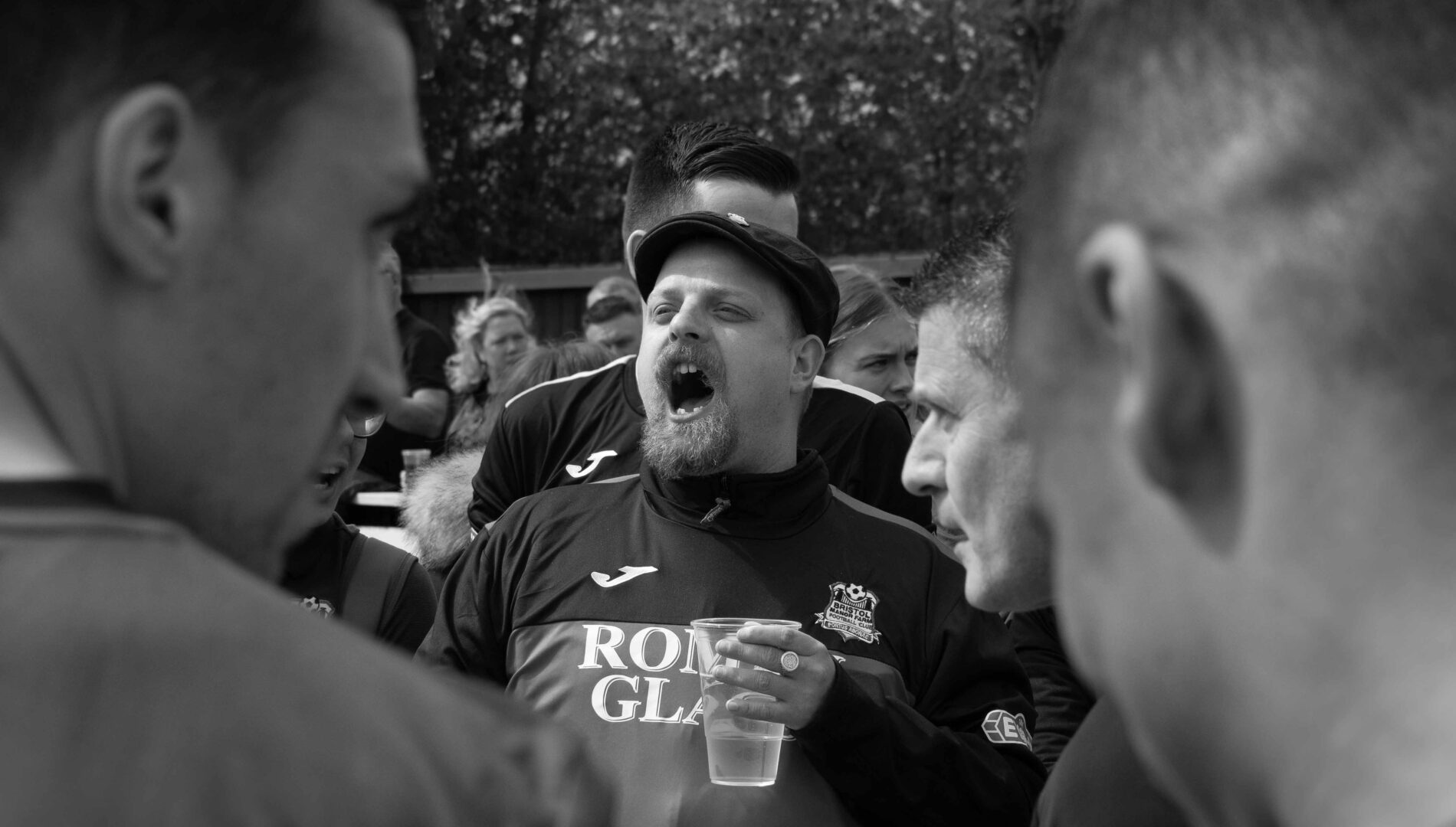 A football fan wearing a flat cap and holding a pint glass cheers, surrounded by other fans.